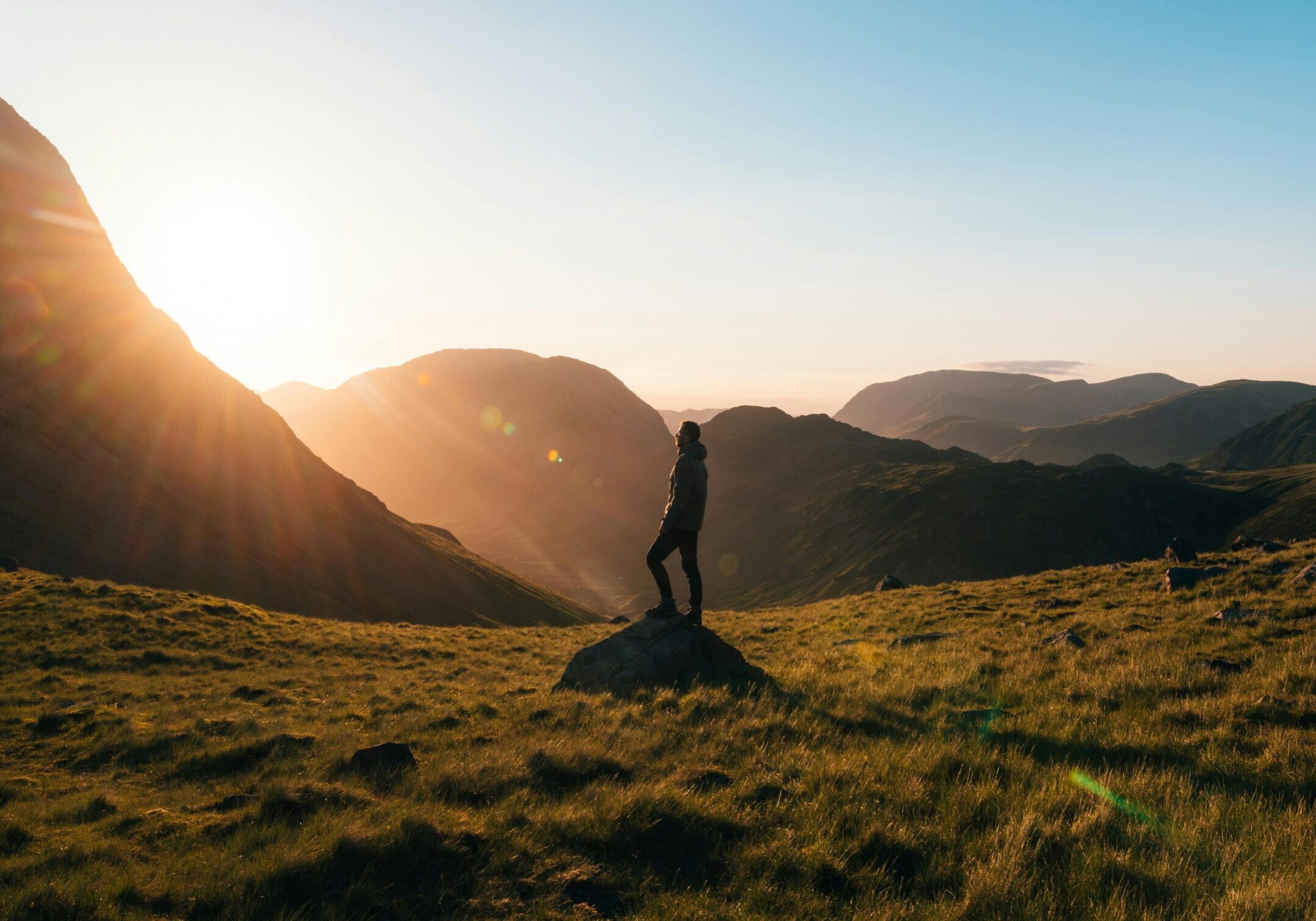 Silhouette Photography of Person Standing on Green Grass in Front of Mountains during Golden Hour